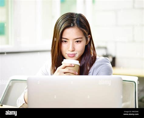 Beautiful Young Asian Business Woman Working In Office Holding A Cup Of Coffee Looking At Laptop