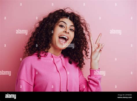 Excited Bi Racial Girl Showing Victory Gesture While Looking At Camera On Pink Background Stock