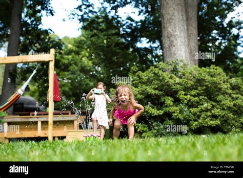 Girl Photographing While Sister Playing With Sprinkler In Backyard