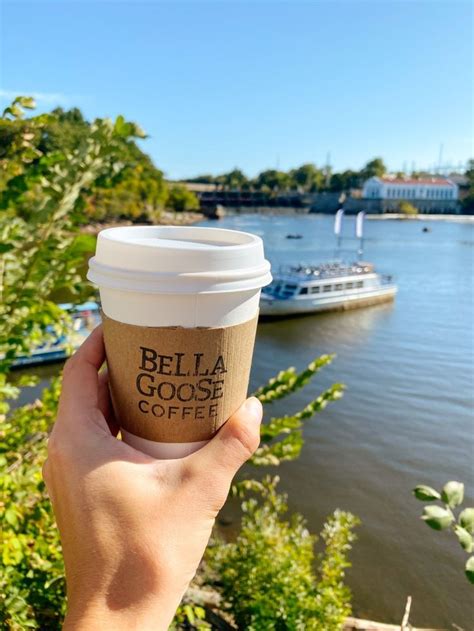 A Person Holding Up A Cup Of Coffee In Front Of A River With Boats On