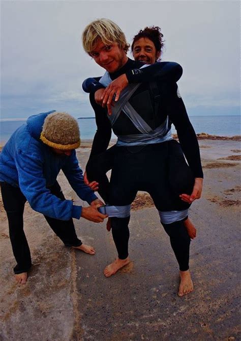 Three People In Wetsuits Standing On The Beach With Their Arms Around