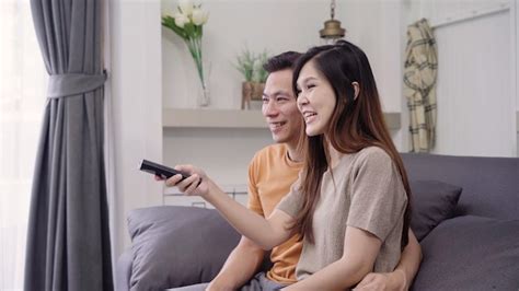 アジアのカップルがテレビを見ていると自宅の居間で暖かいコーヒーを飲む、甘いカップル 無料の写真