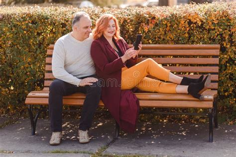 Middle Aged Couple Sit On A Park Bench And Take Selfies On Their Smartphone Stock Image Image