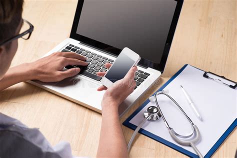 Male Doctor Using A Laptop And Smartphone Sitting At His Desk Stock