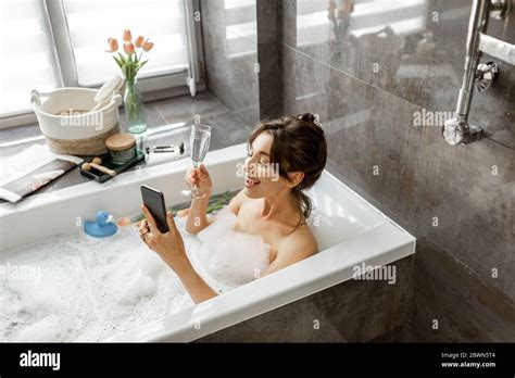 Young Woman Taking A Bath Drinking Sparkling Wine Talking On Phone Lying In Bathtub With Foam