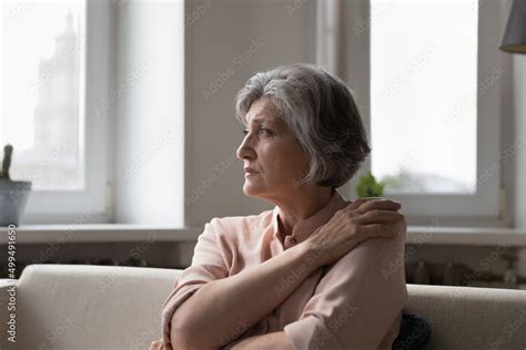 older woman sit on couch in living room staring into distance feeling lonely suffers from