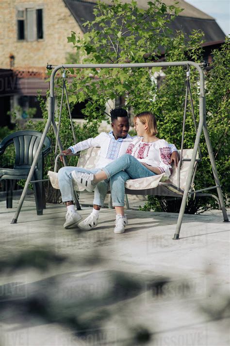 Interracial Couple Sits On Bench Swing In Garden Dressed In Ukrainian