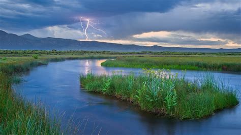 River Between Green Grass Field With A View Of A Lightning And