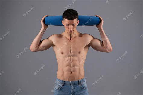 Парень без рубашки с подтянутым торсом держит тренировочный коврик на сером фоне фитнес