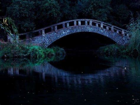 A Stone Bridge Over A Body Of Water At Night With Trees And Bushes In