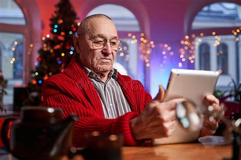 Serious Old Man In Red Sweater Poses Sitting At Table With Tablet Stock