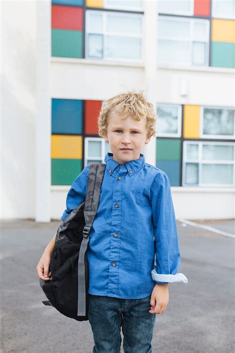 A Blonde Boy Standing In Front Of A School With A Backpack On His