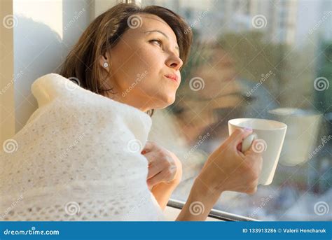 Mature Woman Standing Near The Window With Cup Of Hot Drink Under Warm Knitted Woolen Blanket