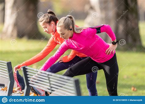 Two Female Athletes Are Warming Up In The Autumn Park Using A Bench To