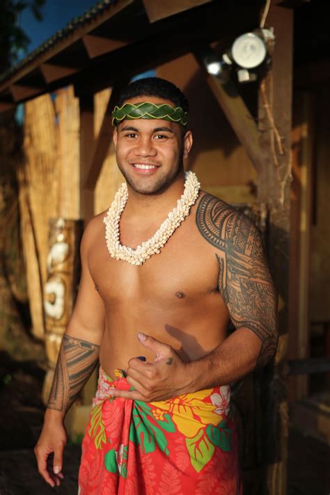 A Man With Tattoos On His Chest Wearing A Red And Green Hawaiian Sarong Standing In Front Of A Hut