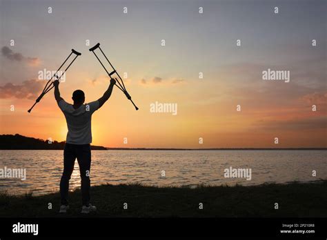 Man Raising Hands With Underarm Crutches Up To Sky Near River At Sunset