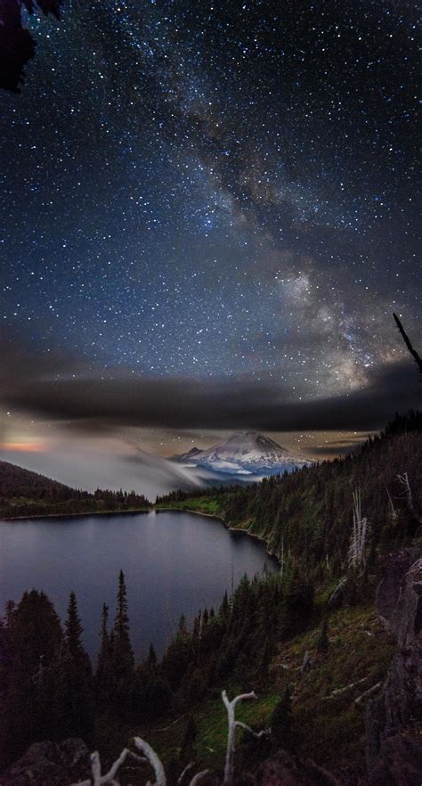The Night Sky Is Filled With Stars And Clouds Above A Lake Surrounded