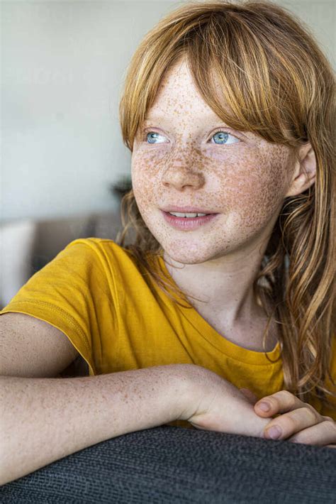Smiling Redhead Girl With Freckles Looking Away While Sitting On Chair At Home Stockphoto
