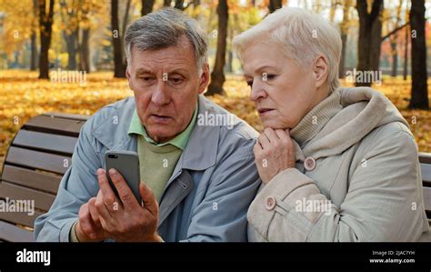 Upset Old Married Caucasian Couple Use Smartphone Sit On Bench In Autumn Park Look At Screen