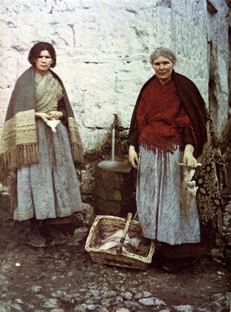 Two Women Standing Next To Each Other In Front Of A Stone Building With A Basket On The Ground