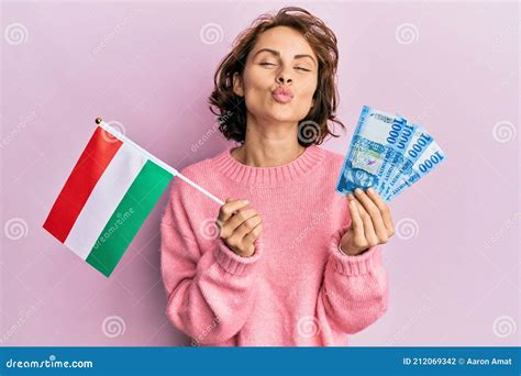 Young Brunette Woman Holding Hungary Flag And Forints Banknotes Looking At The Camera Blowing A