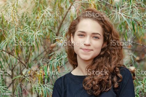 Portrait Of A Girl With Brown Curly Hair On A Background Of Green Bush