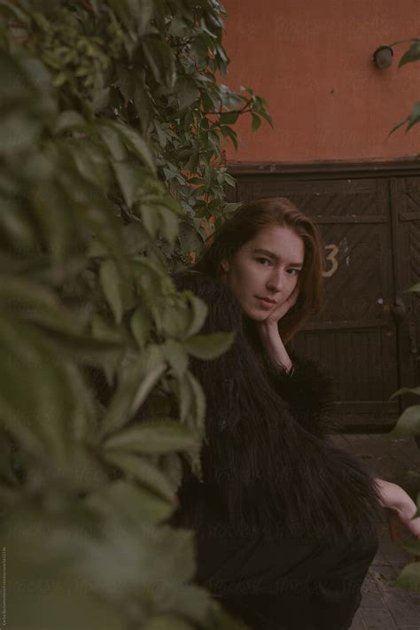 Portrait Of A Redhead Girl With Freckles In A Long Black Dress And A Fur Coat Near The Wall