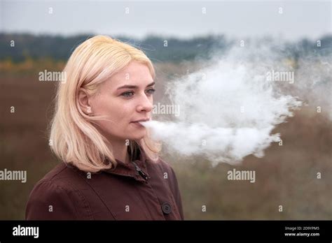 Young Blonde Woman Exhales Cigarette Smoke On Nature In Autumn Stock