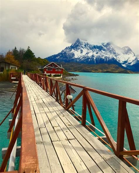 A Wooden Bridge Over Water With Mountains In The Backgroung And Clouds