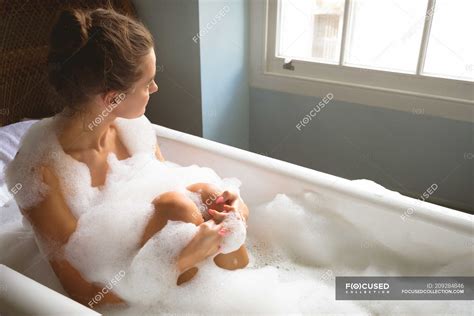 Woman Taking Bath With Foam In Bathtub And Looking Through Window At Home Indoors Wellness