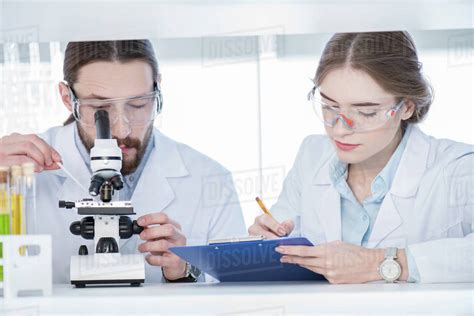 Two Coworkers Chemists Working With Microscope And Taking Notes In