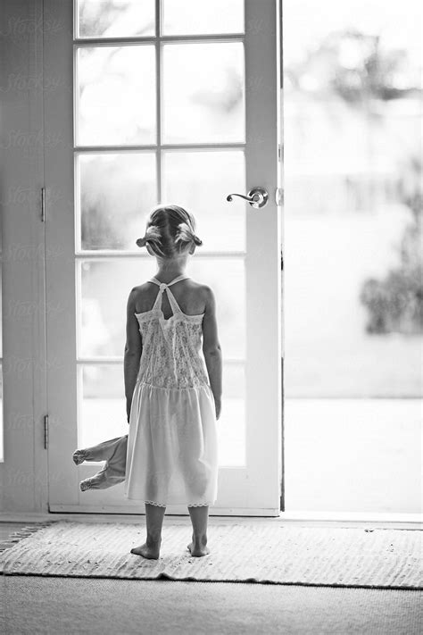 Girl In White Dress Holding Doll Facing French Door By Stocksy
