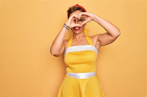 middle age senior pin up woman wearing 50s style retro dress over yellow background doing heart