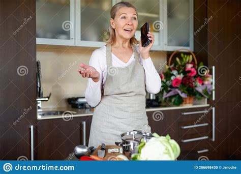 Mature Woman In Kitchen Preparing Food And Holding Smart Phone Stock