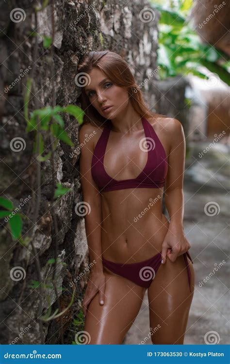 Brunette With Shining Tanned Skin Posing In Burgundy Bikini Near Stone Wall In Tropics Stock