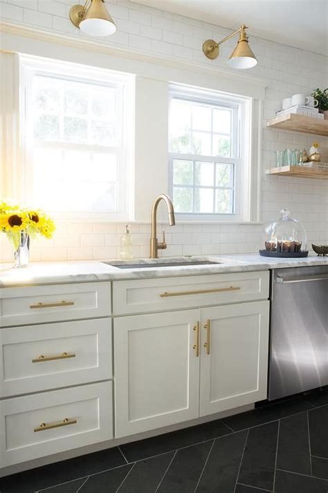 Kitchen features a blue kitchen island topped with black leathered granite countertop and light brown nailhead counter stools, illuminated by bronze and white glass pendants and cream shaker cabinets. White and Gold Kitchen with Schoolhouse Electric Princeton ...