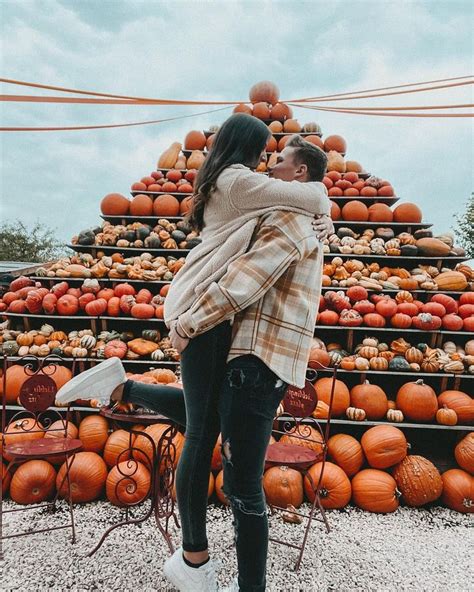 A Man And Woman Standing In Front Of A Pile Of Pumpkins With Their Arms Around Each Other