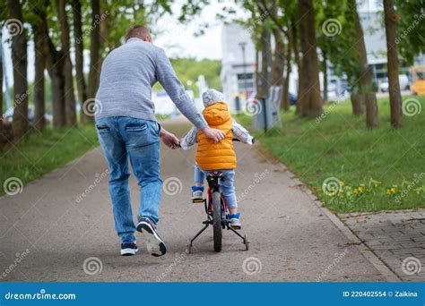 Father Help Son To Ride Bicycle In Summer Park Back View Stock Photo