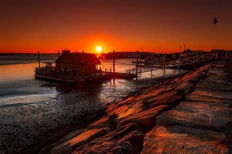 The Sun Is Setting Over Some Water With Rocks In Front Of It And A House