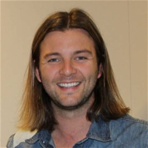 A Man With Long Hair Smiling At The Camera And Wearing A Denim Shirt