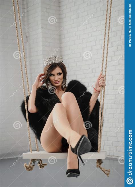 Fashion Woman In Black Fur Coat And Pink Underwear With A Crown On Her Head Ridding On A Swing