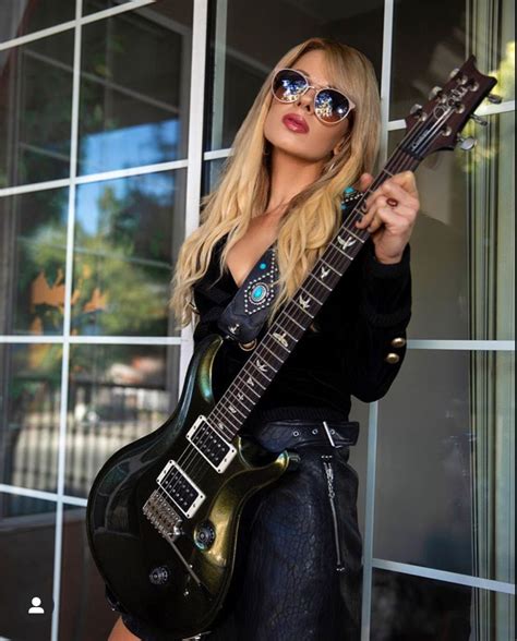 a woman with sunglasses holding a guitar in front of a glass building and posing for the camera