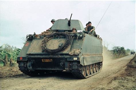 Two Men Are Riding On Top Of An Army Tank In The Middle Of A Dirt Road