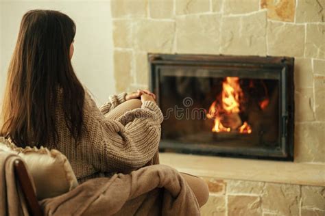 Stylish Woman In Cozy Sweater Warming Up In Chair At Fireplace In