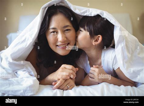 Daughter Kissing Her Mother Under Blanket In Bedroom At Home Stock