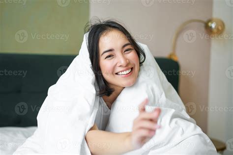 Beautiful asian woman sitting on bed, covered with white duvet, smiling