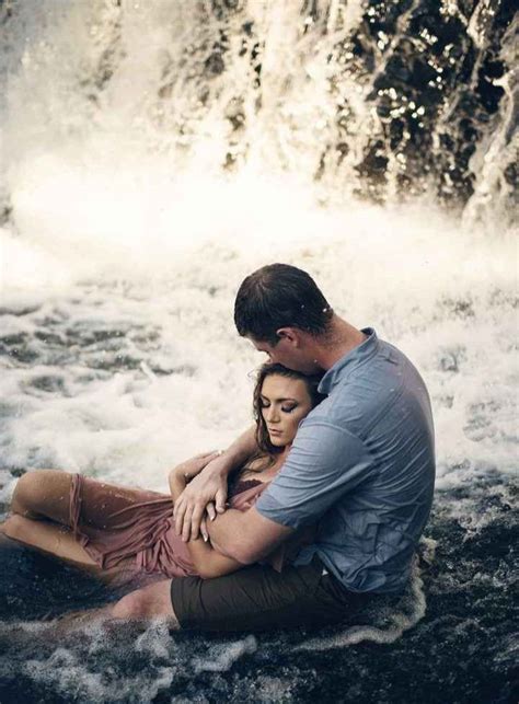 A Man And Woman Sitting On The Ground In Front Of A Waterfall With Their Arms Around Each Other