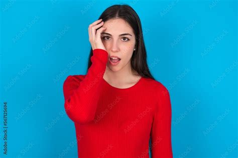 Embarrassed Young Brunette Girl Wearing Red T Shirt Against Blue Wall With Shocked Expression