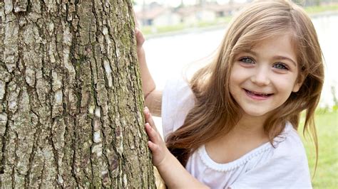 Smiley Cute Little Girl Is Standing Near Tree Trunk Wearing White Dress