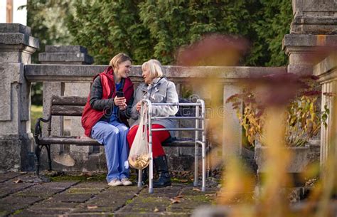 Senior Woman With Walking Frame And Caregiver Outdoors Sitting In Park